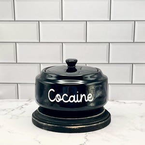 May include: A black ceramic jar with a lid and a knob, sitting on a wooden base. The word "Cocaine" is written in white script on the front of the jar. The background features white subway tile.