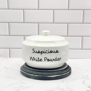 May include: A white ceramic lidded jar with a small knob handle on top. The front of the jar features the black text "Suspicious White Powder" in a simple sans-serif font. The jar sits on a round, distressed black wooden base with visible wear.
