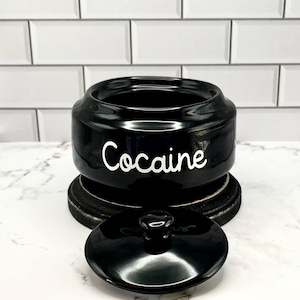 Puede incluir: Un tarro de cer&aacute;mica negro con tapa, sobre una base de madera oscura. La palabra "Cocaine" est&aacute; escrita en blanco en la parte delantera del tarro. La tapa est&aacute; quitada, revelando el interior del tarro.