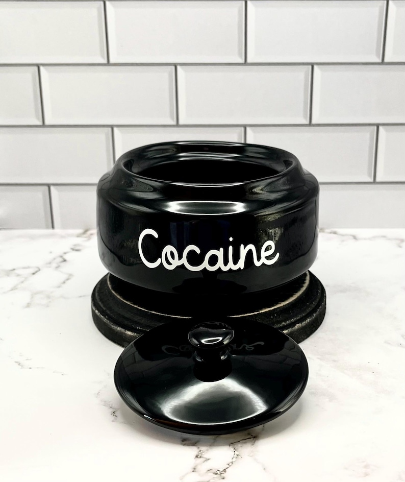 Puede incluir: Un tarro de cer&aacute;mica negro con tapa, sobre una base de madera oscura. La palabra "Cocaine" est&aacute; escrita en blanco en la parte delantera del tarro. La tapa est&aacute; quitada, revelando el interior del tarro.
