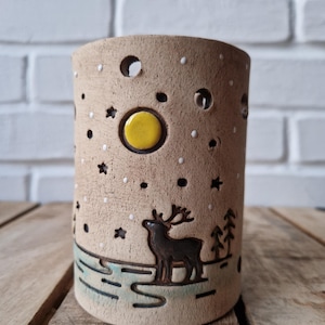 Könnte beinhalten: Ein zylindrischer Kerzenhalter aus Keramik mit einer Winterszene. Der beige Halter zeigt ein Reh, Bäume und ein Monddesign. Das Design umfasst ausgeschnittene Sterne und Punkte, mit einem gelben Mond und einer blau-grünen Landschaft.