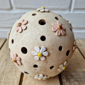 Könnte beinhalten: Ein kugelförmiges Keramikobjekt mit texturierter beiger Oberfläche, das mehrere kreisförmige Aussparungen und applizierte dreidimensionale Blumendekorationen in Rosa- und Weißtönen mit gelben oder braunen Zentren auf der Außenseite aufweist.
