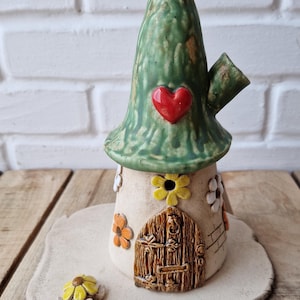 Könnte beinhalten: Eine Keramikhaus-Figur mit einem hohen, texturierten grünen Dach und einem roten Herz. Der beige Sockel hat eine braune Bogentür, gelbe und orangefarbene Blumenakzente sowie eine kleine gelbe Blumenschildkröte auf einer flachen Tonplatte.