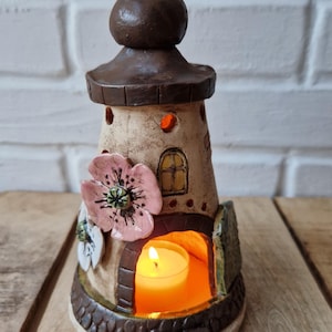 Könnte beinhalten: Ein keramikförmiger Leuchtturm-Kerzenhalter mit braunem Dach und Sockel sowie einem beigefarbenen Körper. Er hat eine rosa Blume und eine brennende Kerze im Inneren. Der Kerzenhalter ist etwa 15 cm hoch.