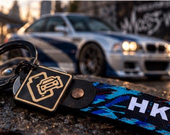Könnte beinhalten: Ein schwarzer Schlüsselanhänger mit einem quadratischen Metallanhänger mit goldenem Auto-Symbol und einem Stoffband mit blau-schwarzem abstraktem Muster. Das Band zeigt den weißen Schriftzug HKS in Großbuchstaben und ist mit einer Kupferniete befestigt.