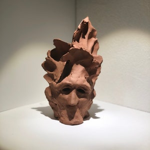 Peut inclure: Une sculpture en argile brune d'un visage avec une surface texturée. Le visage a une large bouche et de grands yeux.