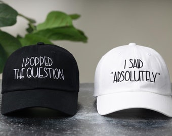 May include: Two baseball caps, one black with white text that says "I popped the question" and one white with black text that says "I said "absolutely""