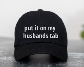 May include: A black baseball cap with the text "put it on my husbands tab" in white.