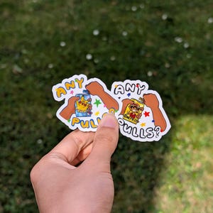 May include: Two stickers held in a hand, featuring cartoon illustrations and text. One sticker has a blue can with the text "ANY PULLS?" and the other has a character from "ONE PIECE" with the text "ANY PULLS?". The stickers have a white border.