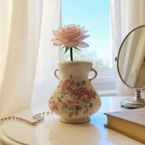 Op de afbeelding: Een kleine, crèmekleurige vaas met twee handvatten en een bloemenontwerp, met een enkele roze roos. De vaas staat op een wit oppervlak, met een parelketting en een vintage spiegel op de achtergrond. De scène wordt verlicht door natuurlijk licht.