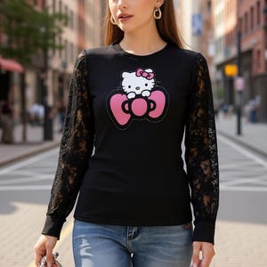 May include: Black long-sleeve shirt with a Hello Kitty graphic. The shirt features a round neck and black lace sleeves. The graphic shows Hello Kitty with a pink bow on her head, centered on a pink bow.