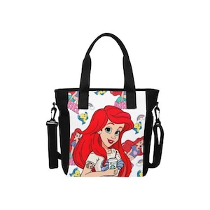 Puede incluir: Bolso tote negro con una colorida ilustraci&oacute;n de Ariel de La Sirenita. El dise&ntilde;o incluye a Ariel sosteniendo una taza de caf&eacute;, con un tatuaje en el brazo. El bolso tiene asas negras y una correa para el hombro desmontable.