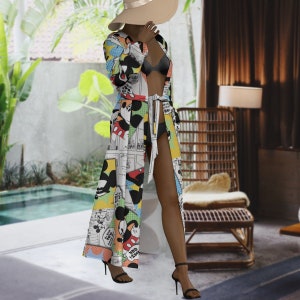 May include: A long, open-front robe featuring a colorful Mickey Mouse comic strip pattern. The robe has a tie waist and is paired with a black bikini and high-heeled sandals. A wide-brimmed hat completes the look.