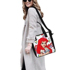 Puede incluir: Bolso tote negro y blanco con una colorida ilustraci&oacute;n de Ariel de La Sirenita. El bolso tiene una correa para el hombro y asas negras. El dise&ntilde;o incluye a Ariel con cabello rojo y una camisa blanca, sosteniendo una bebida.