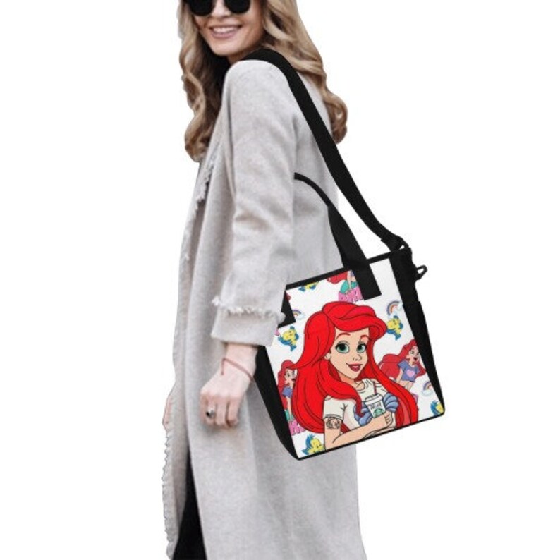 Puede incluir: Bolso tote negro y blanco con una colorida ilustraci&oacute;n de Ariel de La Sirenita. El bolso tiene una correa para el hombro y asas negras. El dise&ntilde;o incluye a Ariel con cabello rojo y una camisa blanca, sosteniendo una bebida.