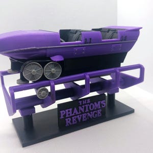May include: A purple roller coaster car model with black seats and grey wheels, mounted on a purple track section. A black rectangular sign in front features the purple text THE PHANTOM'S REVENGE. The entire assembly sits on a black base.