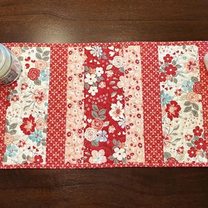 May include: A red floral table runner with a patchwork design. The runner features alternating panels of floral and gingham patterns. A candle in a glass jar and a white swan vase with flowers are on the runner.