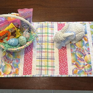 May include: A colorful Easter table runner with a basket of treats and a decorative lamb. The runner features pastel eggs, bunnies, and plaid patterns. The basket contains candy and decorated eggs, perfect for Easter celebrations.