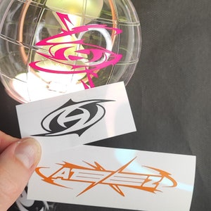 Könnte beinhalten: Zwei weiße Aufkleber mit schwarzen und orangen Logos. Der erste Aufkleber hat ein schwarzes Logo mit den Buchstaben "A" und "Z", die ineinander verschlungen sind. Der zweite Aufkleber hat ein oranges Logo mit dem Text "ATEZZ" in einer stilisierten Schrift.