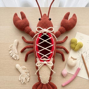 Puede incluir: Un juguete de peluche de langosta hecho a ganchillo en color óxido con una abertura central atada con un cordón blanco, dos pinzas grandes y varias patas. Junto a él hay pequeños accesorios de ganchillo, incluyendo dos aletas color crema, un grupo amarillo y una pieza rosa.