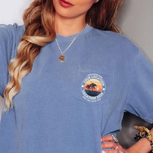 May include: A blue jean shirt with a pocket featuring a graphic of palm trees and a sunset with the text "Your Location Vacation 2024".