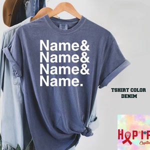 May include: A denim blue t-shirt with the text "Name & Name & Name & Name." printed in white. The t-shirt is displayed on a hanger with a denim shirt and a plant in the background.