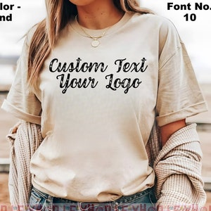 May include: A sand colored t-shirt with the text "Custom Text Your Logo" in a black font. The font is a cursive script style.