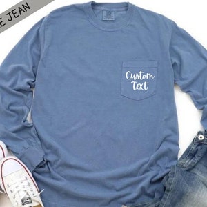 以下が含まれることがあります： 左側にポケットが付いた青い長袖Tシャツ。ポケットには「Custom Text」という白い文字が書かれています。シャツは白い背景に白いスニーカーと青いジーンズを合わせています。