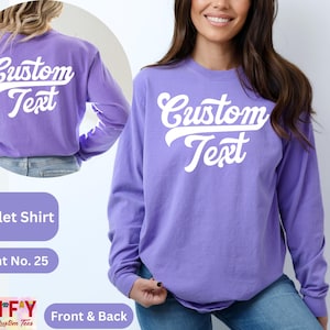 Pode incluir: Uma camisa de manga comprida roxa com o texto "Custom Text" em uma fonte retro branca na frente e nas costas. A camisa &eacute; mostrada em uma modelo.