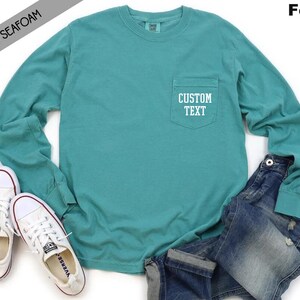 以下が含まれることがあります： 左側にポケットが付いたターコイズブルーの長袖Tシャツ。ポケットには白い文字で「CUSTOM TEXT」と書いてあります。シャツは白い背景に白いスニーカーと青いジーンズを合わせています。