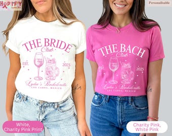 May include: Two white t-shirts with pink text and graphics. The text on the left shirt reads "The Bride Club Est. 2025 Lydia's Bachelorette Los Cabos, Mexico". The text on the right shirt reads "The Bach Club Est. 2025 Lydia's Bachelorette Los Cabos, Mexico".