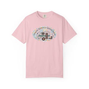 May include: A light pink t-shirt featuring an oval design with a vintage camper illustration. The camper is decorated with a floral pattern and the text "Camping, Stitching & Quilting Retreat" is arched around the design.