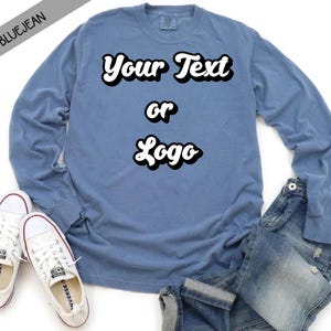 May include: A blue long-sleeve t-shirt with the text "Your Text or Logo" in a retro font. The shirt is shown on a model wearing jeans and white sneakers.