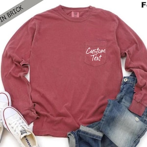 以下が含まれることがあります： 左側にポケットが付いた、レンガ色の長袖Tシャツ。ポケットには白い文字で「Custom Text」と書かれている。モデルは青いジーンズと白いスニーカーを履いている