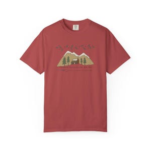 May include: A brick red short-sleeve t-shirt featuring a graphic of a cabin nestled in front of snow-capped mountains and pine trees. Above the scene are green holly sprigs, and below is the text "Christmas blessings in the mountains".