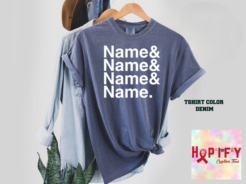 May include: A denim blue t-shirt with the text "Name & Name & Name & Name." printed in white. The t-shirt is displayed on a hanger with a denim shirt and a plant in the background.
