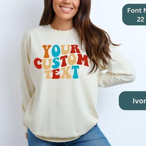 May include: Ivory long-sleeve sweatshirt with the text "YOUR CUSTOM TEXT" in a retro rainbow font. Font No. 22.