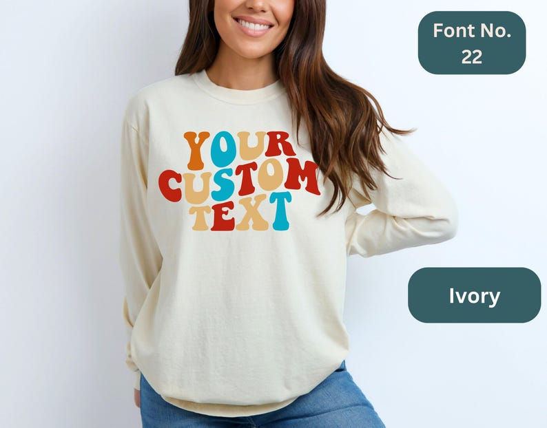 May include: Ivory long-sleeve sweatshirt with the text "YOUR CUSTOM TEXT" in a retro rainbow font. Font No. 22.