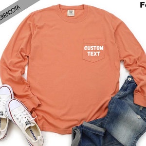 以下が含まれることがあります： 左側にポケットが付いたテラコッタ色の長袖Tシャツ。ポケットには白い文字で「CUSTOM TEXT」と書いてあります。白い背景に白いスニーカーと青いジーンズを合わせたコーディネートで撮影されています。
