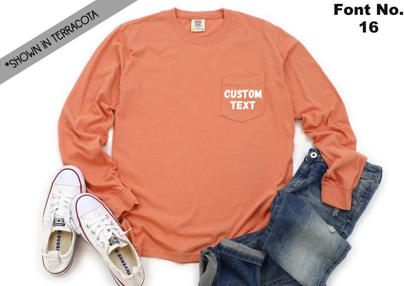 以下が含まれることがあります： 左側にポケットが付いたテラコッタ色の長袖Tシャツ。ポケットには白い文字で「CUSTOM TEXT」と書いてあります。白い背景に白いスニーカーと青いジーンズを合わせたコーディネートで撮影されています。