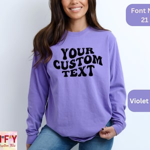 May include: A purple long-sleeve shirt with the text "YOUR CUSTOM TEXT" in a retro font. The shirt is a blank canvas for personalization.