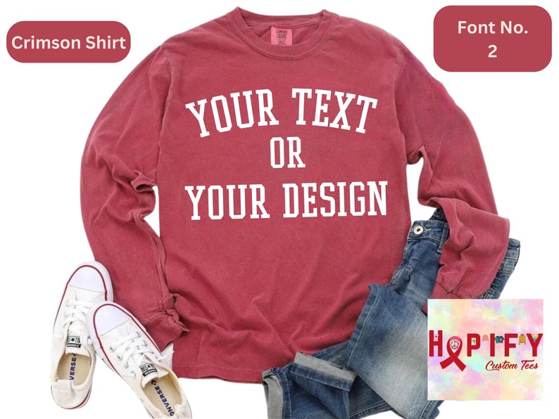 May include: A crimson long-sleeved t-shirt with the text "YOUR TEXT OR YOUR DESIGN" in white. The shirt is laid flat on a surface with a pair of white sneakers and blue jeans.