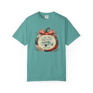 May include: A teal short-sleeve t-shirt featuring a central graphic of a pumpkin with a red and white checkered bow. The pumpkin contains a camper illustration and the text Campin' & Stitchin' Cross Stitch Retreat Fall 2026.