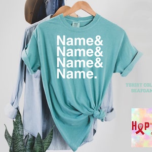 May include: A turquoise blue t-shirt with the text "Name & Name & Name & Name." printed in white. The t-shirt is on a hanger and has a knotted bottom. The text "T-shirt Colour Seafoam" is printed below the shirt.