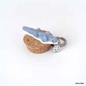 May include: A light blue alligator keychain with a white base, attached to a silver keyring with a smiling face charm. The keychain rests on a cork. The design is simple and playful.
