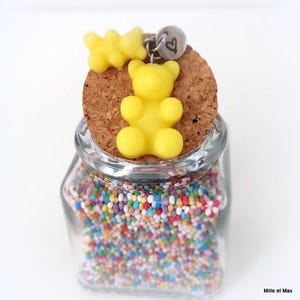 May include: A clear glass jar filled with colorful sprinkles, topped with a cork stopper. A yellow gummy bear charm and a small silver charm with a heart hang from the cork. The jar is labeled "Mille et Max".