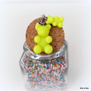 May include: A clear glass jar filled with colorful sprinkles, topped with a cork stopper. A bright yellow gummy bear charm and a smaller gummy bear are attached to the stopper with a silver-colored chain. The jar is set against a white background.