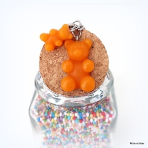 May include: A clear glass jar filled with colorful sprinkles, topped with a cork and a bright orange gummy bear keychain. A smaller gummy bear shape is attached to the keychain. The jar is labeled "Mille et Max".