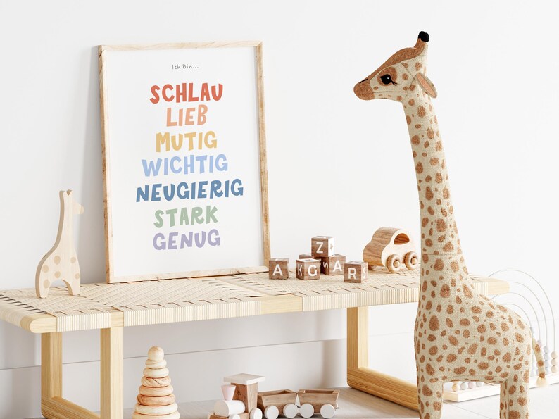 May include: A colorful print with the words "Schlau, Lieb, Mutig, Wichtig, Neugierig, Stark, Genug" in a rainbow color scheme. The print is framed and hanging on a white wall. There is a wooden giraffe toy on a wooden bench in front of the print.