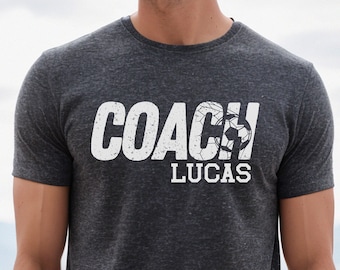 Könnte beinhalten: Ein dunkelgraues T-Shirt mit einem weißen Aufdruck, der "COACH" mit einem Fußball im "O" und "LUCAS" darunter sagt.